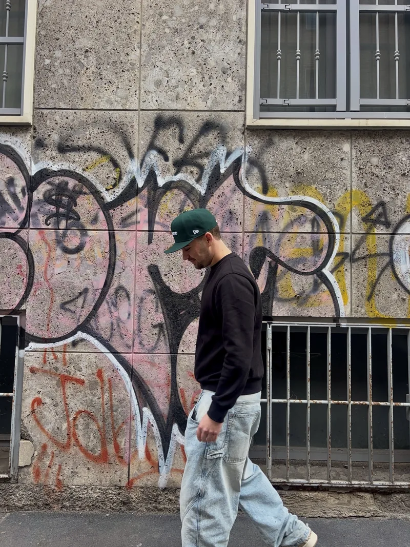 Valentín in a dark sweater and green cap walking past a graffiti wall.